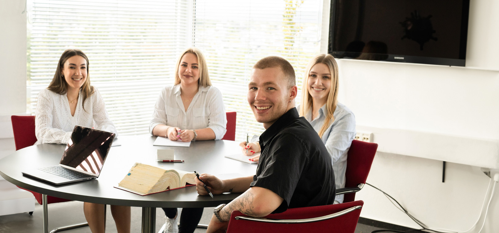 4 junge Mitarbeiter sitzen bei einer Besprechung an einem Tisch, lächeln und blicken Richtung Kamera.